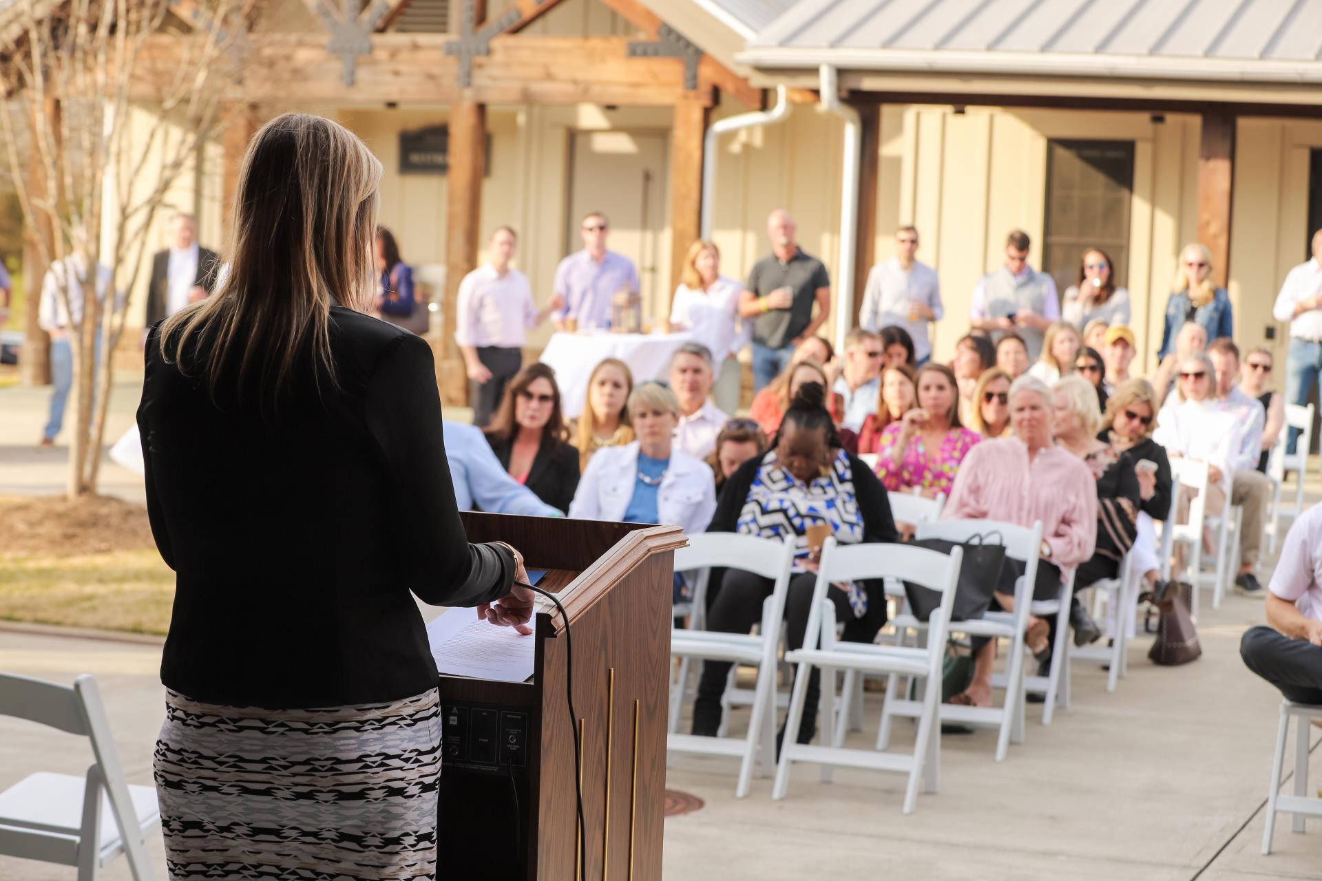 Back of woman standing at podium with view of audience sitting in white chairs in courtyard